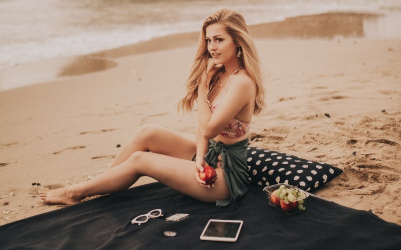 A girl relaxing on the beach while traveling with a tablet and also holding an apple.