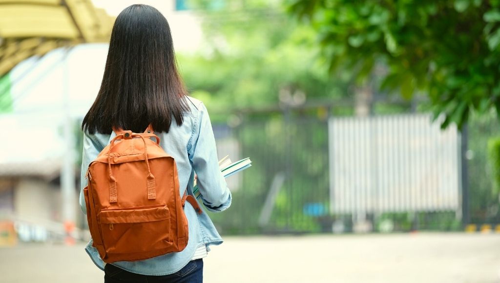 Back of student girl holding books and carry school Daypack while walking in school campus background