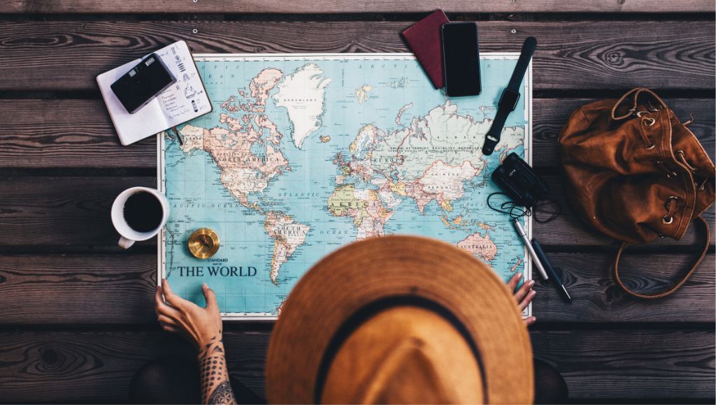 Tourist wearing brown hat looking at the world map and planning vacation using world map and compass along with other travel accessories.