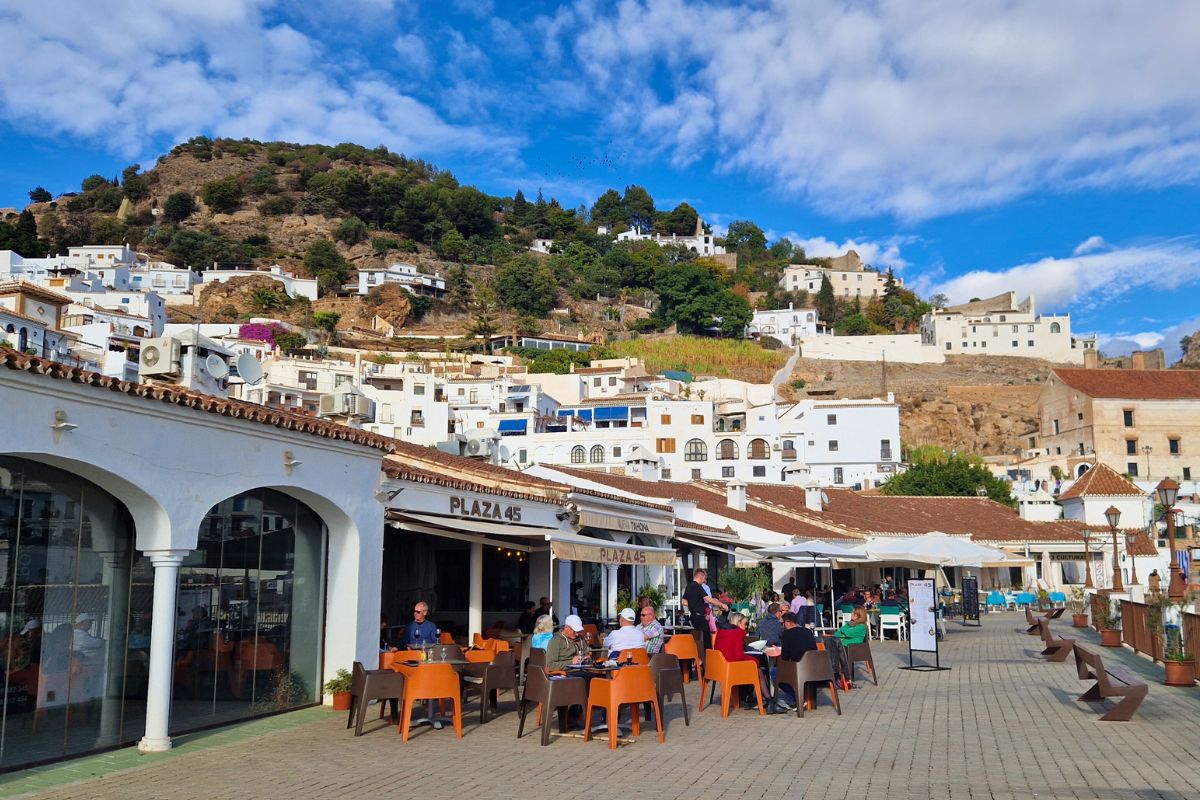 Looking past resturants and people eating in the Plaza of 3 Cultures up to the Andalusian white village Frigiliana