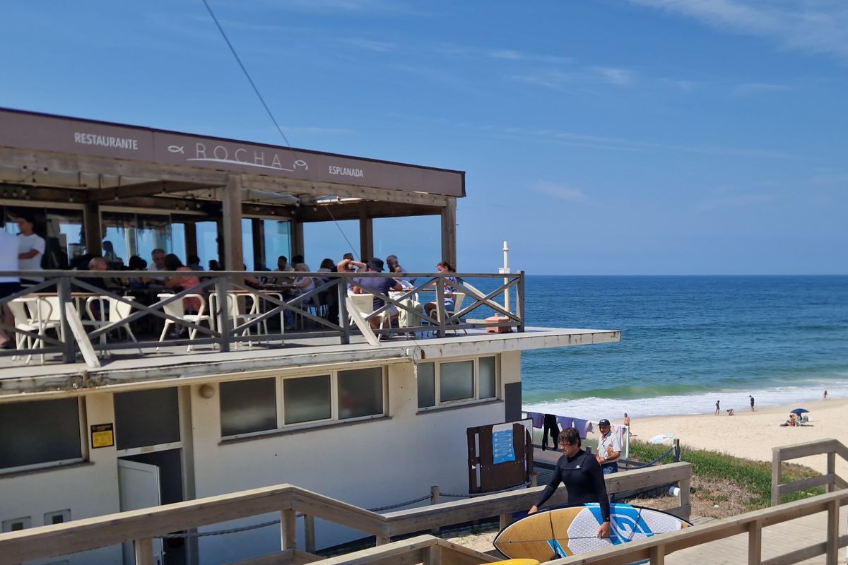 Looking at Restaurante A Rocha with blue skies and ocean the view in the background as a surfer walks past in the foreground